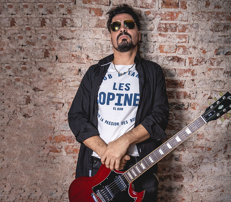 Beto Aguilar con una guitarra roja sobre una pared de ladrillos rustica